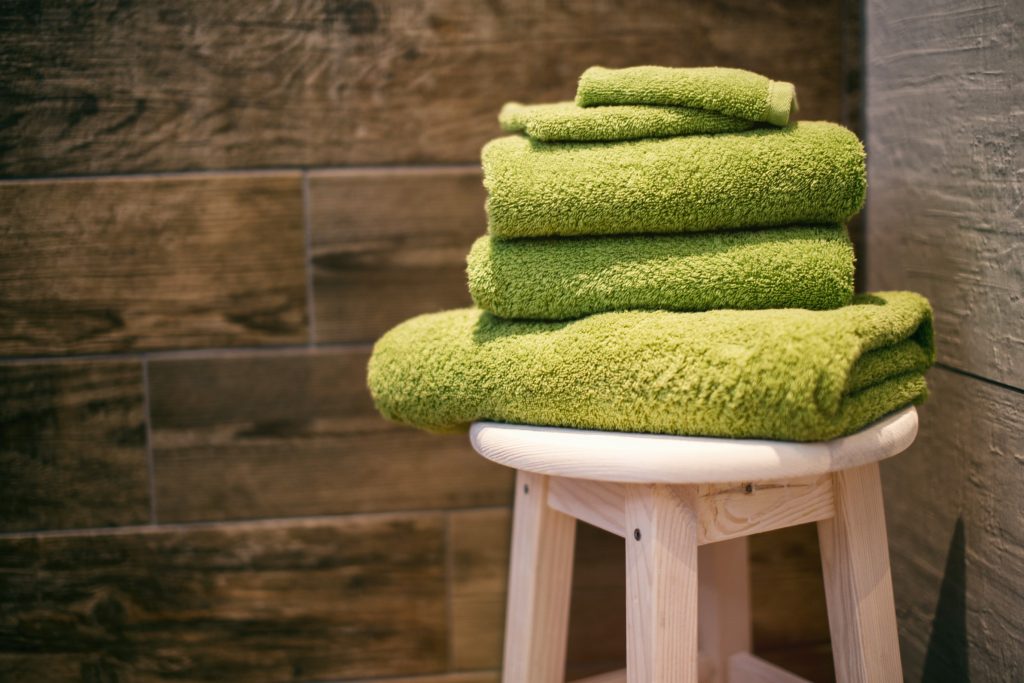 Stack of fluffy lime green bath towels sitting on a wooden stool in a shower with brown wood like rectangular tiles.